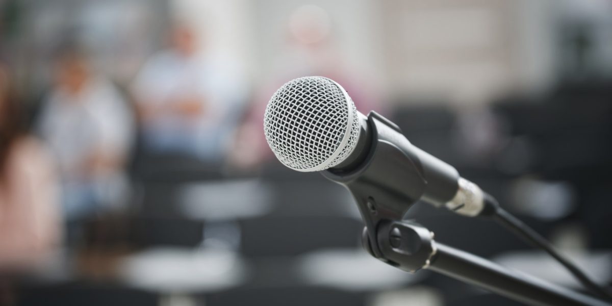 A microphone with a metal grill on a black stand against the background of people and the auditorium or classroom. Public speaking concept. Shallow depth of field. Selective focusing. Macro